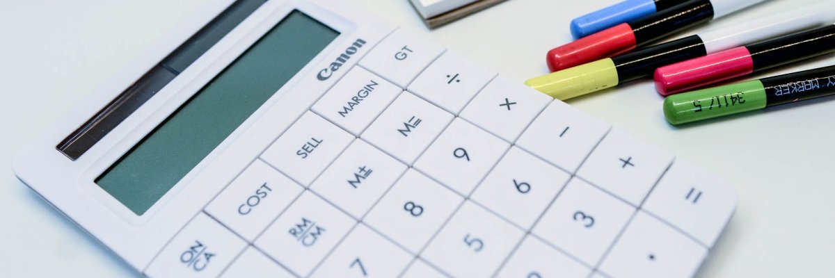 A calculator, glasses, a pad and many pens lie on a table.