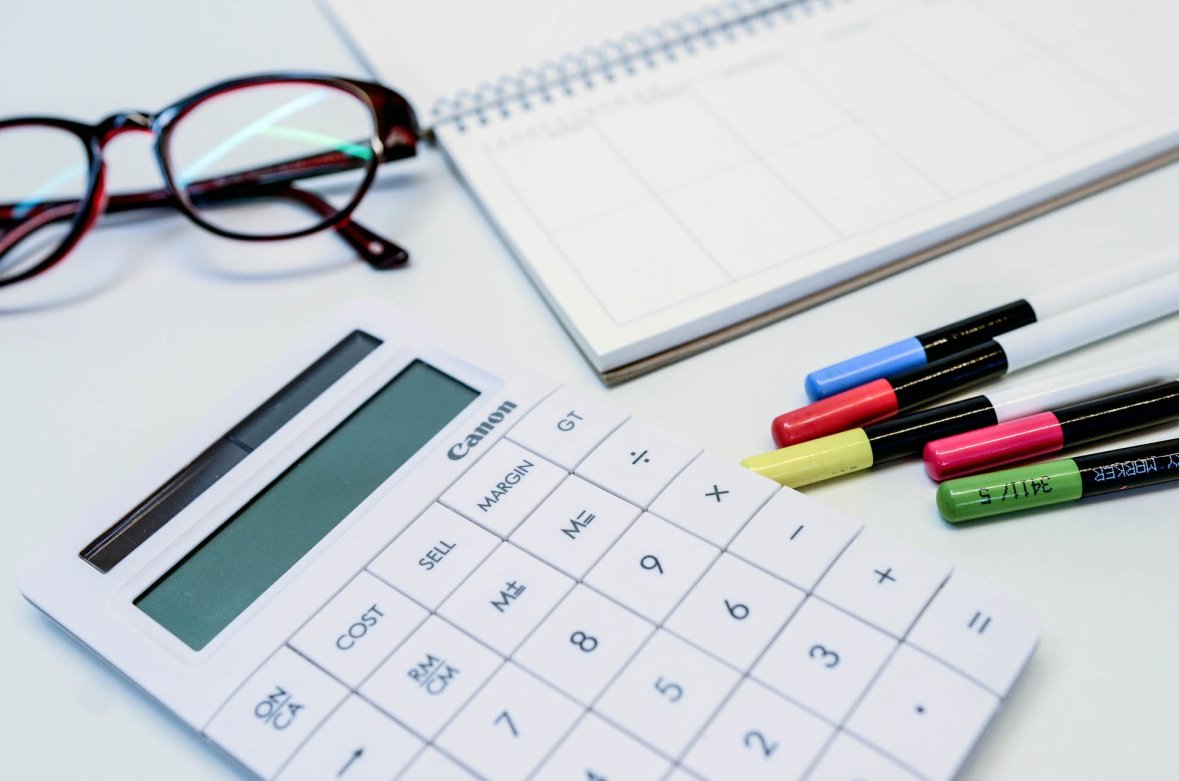 A calculator, glasses, a pad and many pens lie on a table.