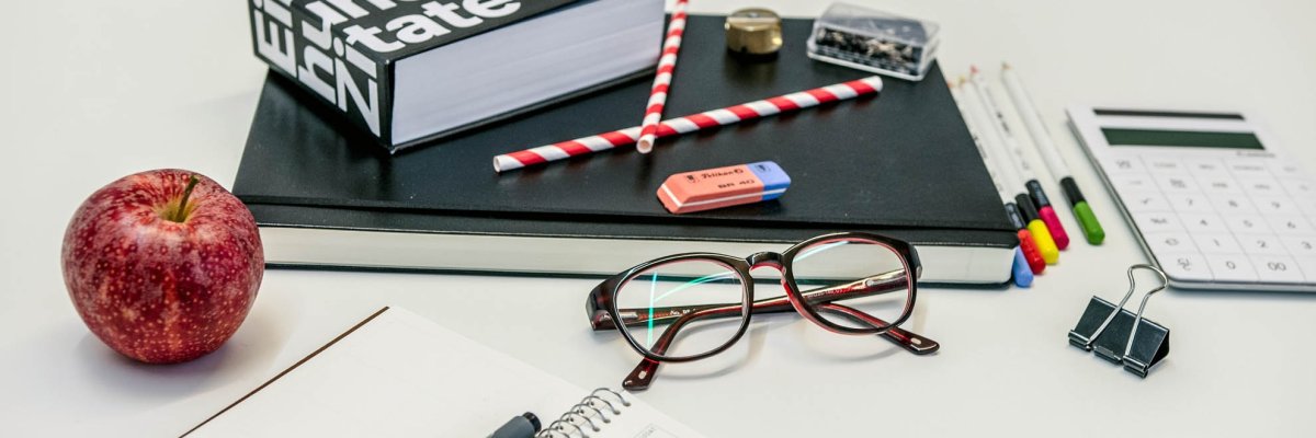 A calculator, a pair of glasses, a notepad, an apple, and many other office supplies lie on a table.
