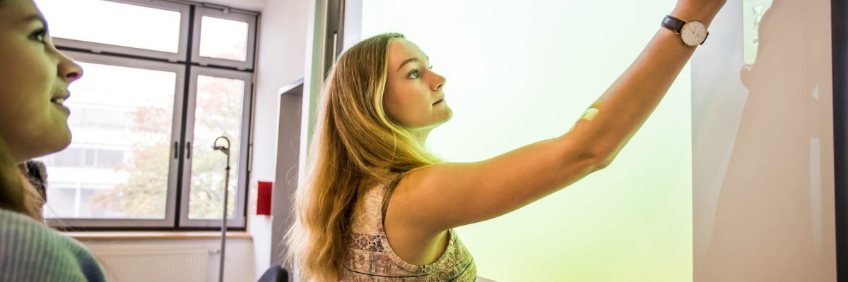 One pupil operates a smartboard, another pupil watches her do it