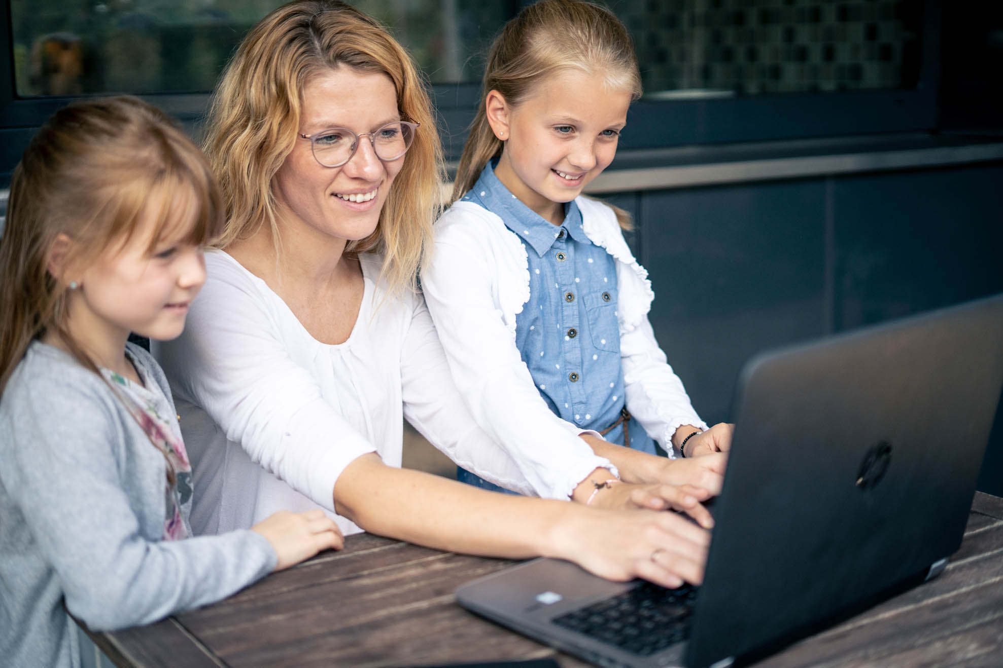 A woman sits at a laptop, two girls stand next to it and look at the screen