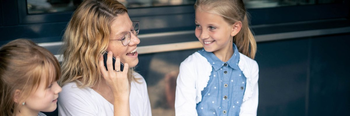 A woman sits on the phone in front of a laptop, two girls stand next to her and look at her.