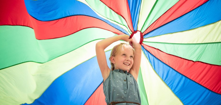 A girl stands under a colourful play parachute and holds it high