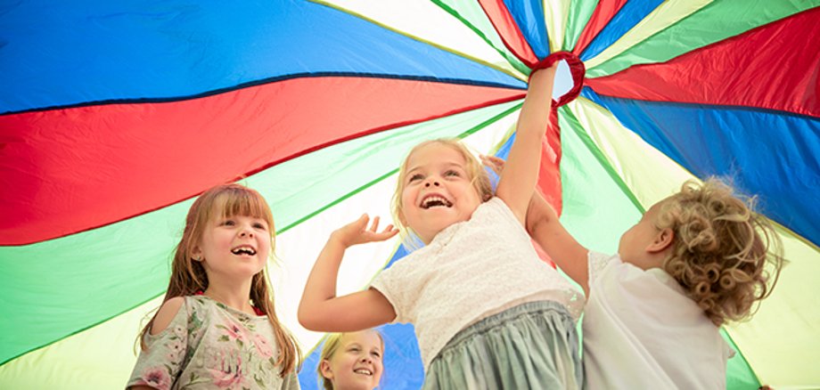 Four children stand under a colourful play parachute and hold it high