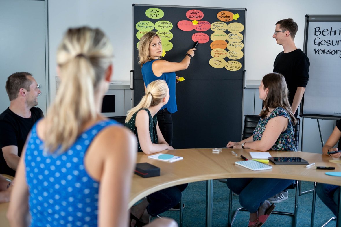 Five people listen to a young woman explaining something on a pin board with slips of paper