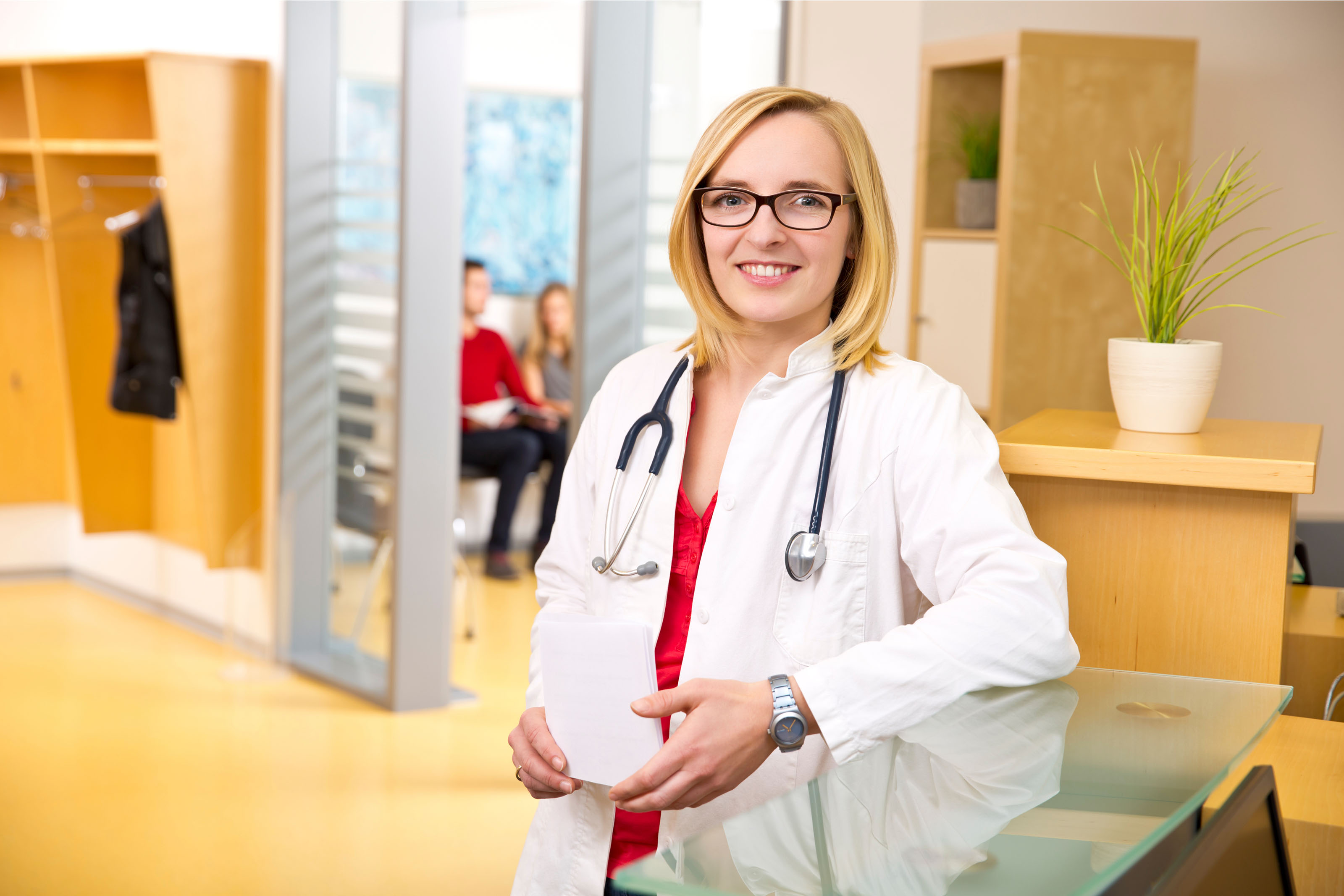 Smiling woman in a doctor's coat with stethoscope around her neck at the reception of a doctor's surgery