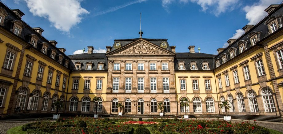 Multi-storey castle building with yellow stones and grey roof and flower border in the inner courtyard