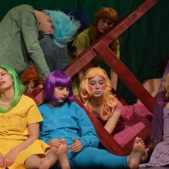 Four colorfully dressed teenagers with colorful wigs sit on the floor in front of a tilted giant chair