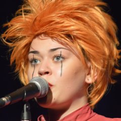 Portrait of a human puppet with red hair speaking into a microphone