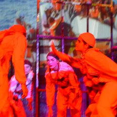 Red-lit harlequin people standing loosely in a circle, leaning into the centre