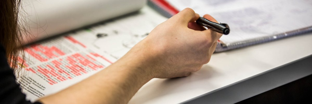 یک دانش‌آموز دختر در حال خواندن از یک کتاب درسی باز است که روی میزی در مقابل او قرار دارد.