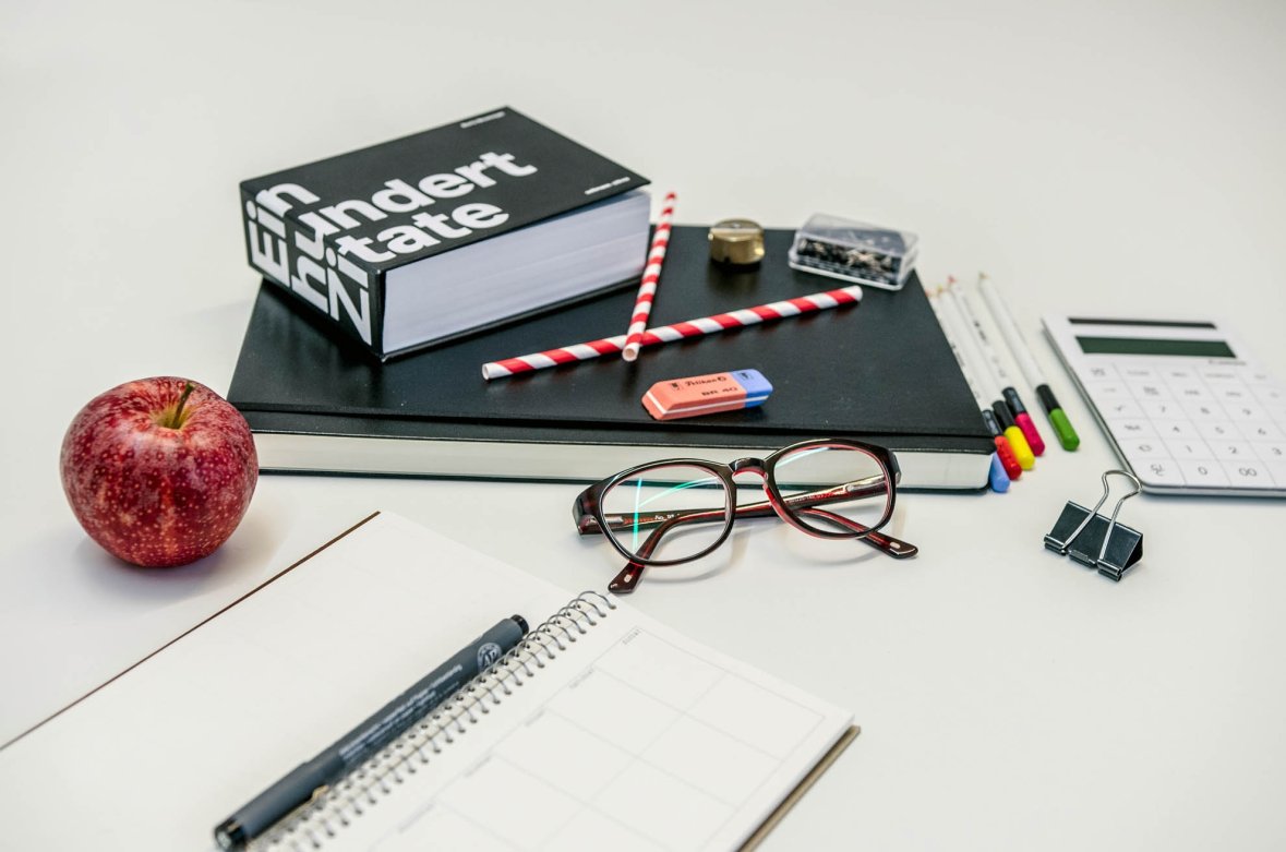 Bir hesap makinesi, bir gözlük, bir not defteri, bir elma ve diğer birçok ofis malzemesi bir masanın üzerinde durmaktadır.