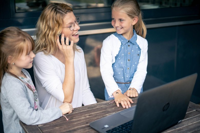 Жінка сидить перед ноутбуком і розмовляє по телефону, дві дівчини стоять поруч і дивляться на неї.