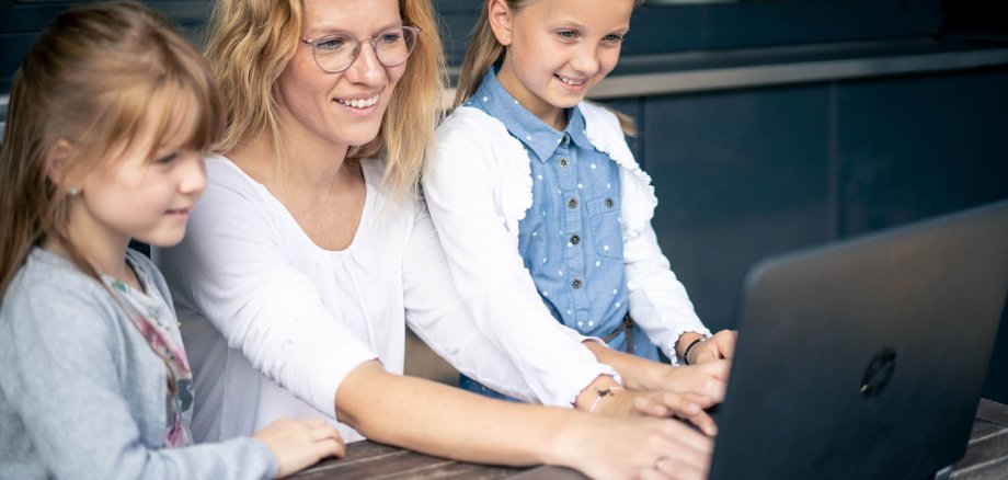 Familie am Computer Eine Frau sitzt an einem Laptop, zwei Mädchen stehen daneben und schauen auf den Bildschirm