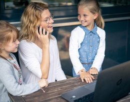 Eine Frau sitzt telefonierend vor einem Laptop, zwei Mädchen stehen daneben und schauen sie an.