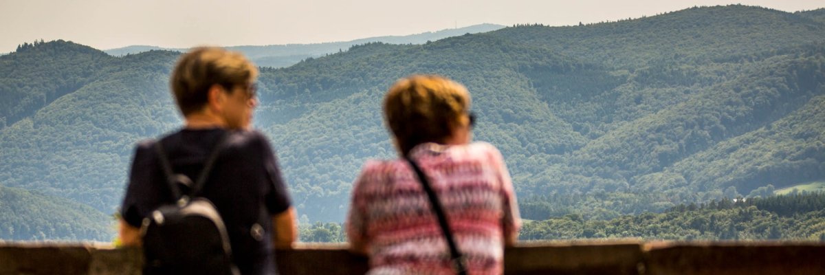 Zwei Frauen in Rückansicht an einer Steinbrüstung mit Blick auf ein Waldgebiet