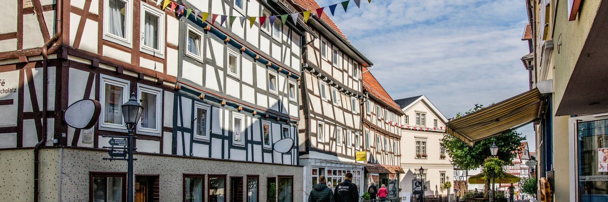 Fachwerkhäuserzeile mit Menschen auf der Straße davor und einer Fahnengirlande auf Höhe des 2. Stockwerks