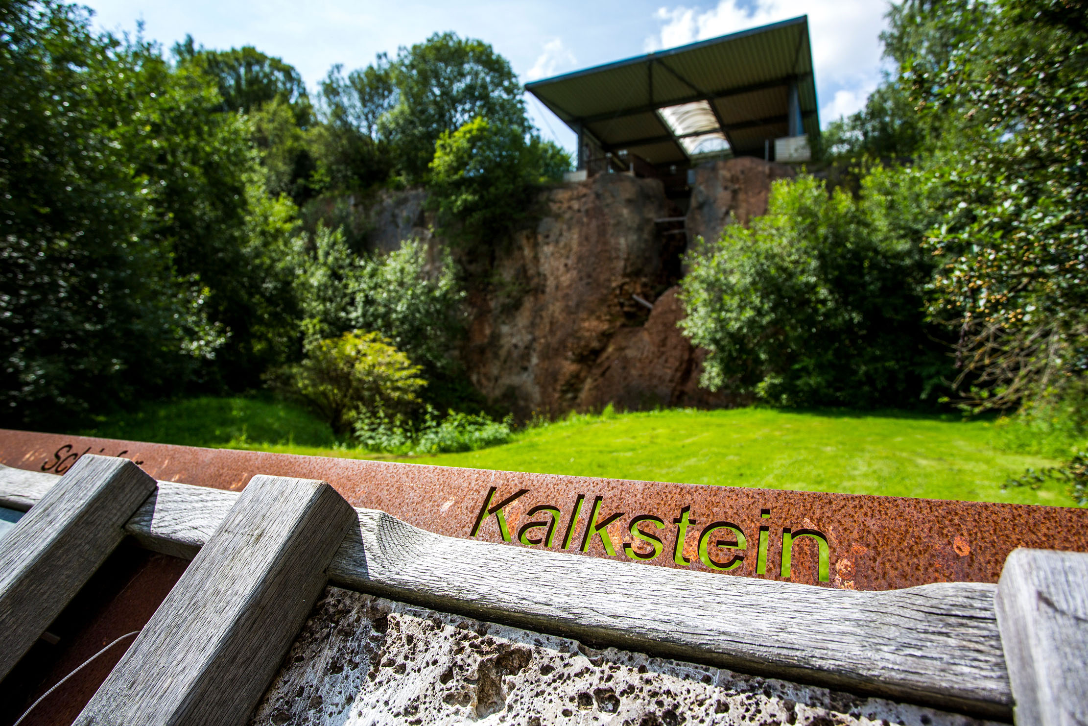 Korbacher Spalte Schriftzug Kalkstein vor einer überdachten Felsspalte mit Büschen und Bäumen umgeben
