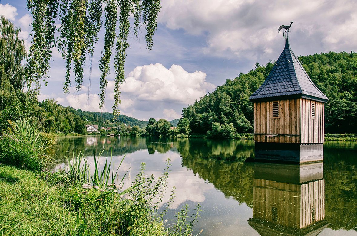 Kirchturm mit Wetterhahn in einem Gewässer umrahmt von Wald und Grünstreifen