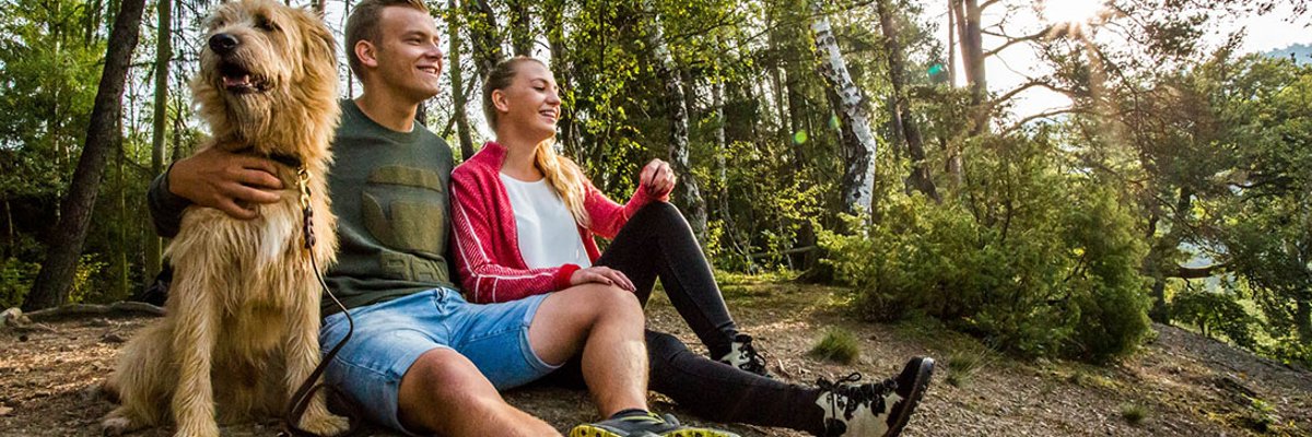 junges Ehepaar mit Hund sitzt auf kargem Waldboden mit Laubbäumen im Hintergrund