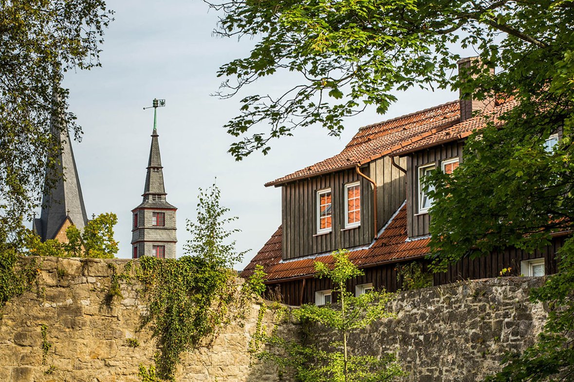 Kirchturmspitze und Turmspitze hinter einer Steinmauer, rechts ragt ein Hausdach über die Mauer hinaus